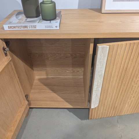 A light oak wood sideboard cabinet with a ribbed fluted front door and an open door revealing a storage compartment. A book and two green vases sit on top, and the cabinet is shown on a gray concrete floor.