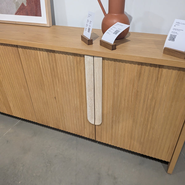 A modern wooden sideboard or credenza with a fluted vertical grain finish, featuring two light-colored vertical handle inserts. A terracotta pitcher and price tags sit on top against a plain light gray wall.