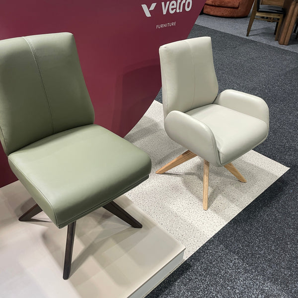 Two modern upholstered lounge chairs displayed in a showroom. One chair is off-white with light wood legs, and the other is sage green with dark metal legs.