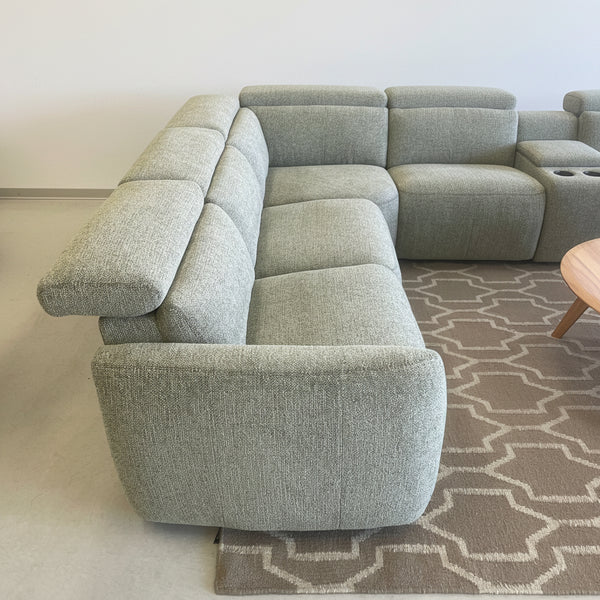 A light sage green bouclé sectional sofa with a right-facing armrest and built-in cup holders, shown in a furniture showroom on a patterned rug.