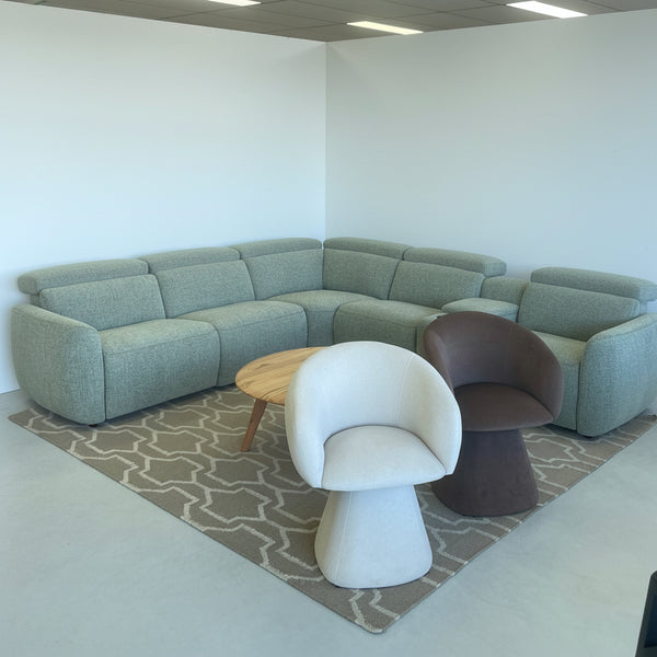 A large U-shaped sectional sofa in a textured sage green fabric, styled with a brown patterned rug, a round wooden coffee table, a modern white molded chair, and a dark brown accent chair in a furniture showroom.