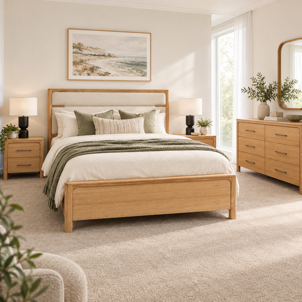A light oak wood bed frame with a upholstered headboard is shown in a bright, modern bedroom setting. The bed is styled with white bedding, green accent pillows, and a green throw blanket, with matching wood nightstands, a dresser, and a framed coastal painting on the wall.