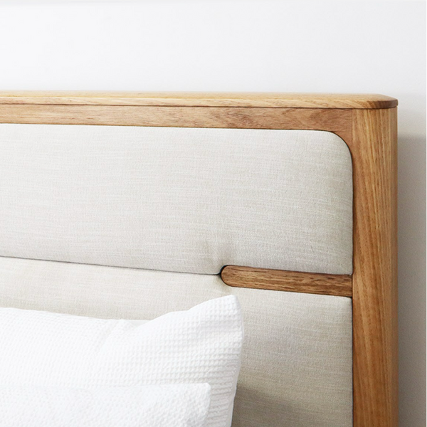 Close-up of a wooden bed headboard with light beige upholstered panels and a white pillow against a plain white wall.
