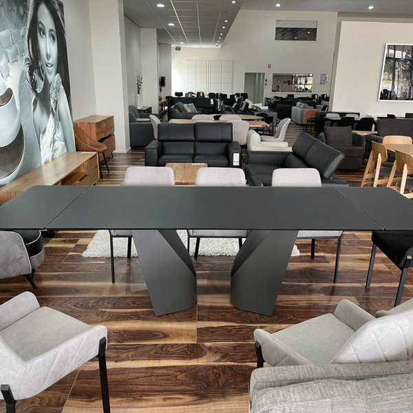 A modern dining table with a matte black rectangular top and sculptural twisted pedestal bases, surrounded by contemporary dining chairs in black, gray, and light gray, set in a furniture showroom.