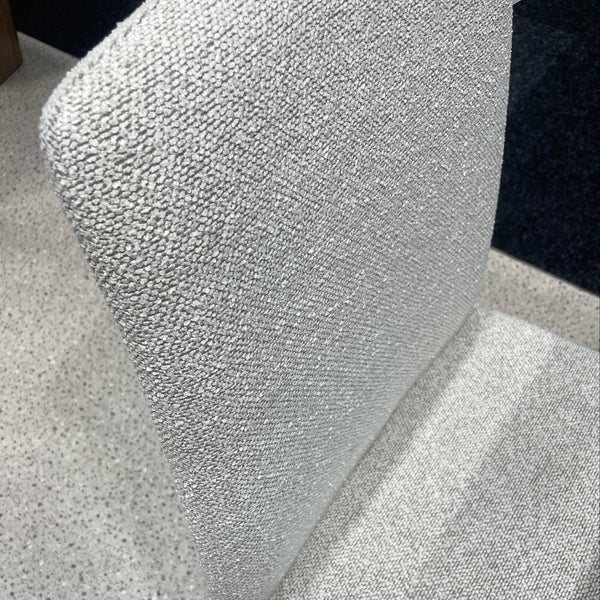 Close-up of a light gray upholstered chair or cushion with a textured woven fabric, shown against a speckled gray floor and dark background.