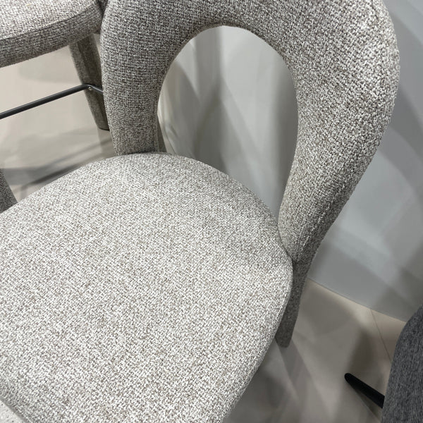 A close-up of a modern upholstered chair with a curved, looped backrest and a rounded seat cushion, both covered in a textured light gray woven fabric.