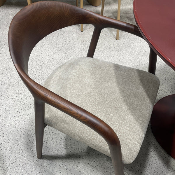 A modern dining chair with a curved dark brown wooden frame and a light beige textured fabric seat cushion, shown on a speckled gray floor.