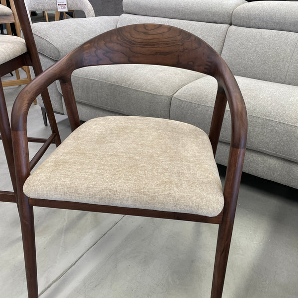 A modern dining chair with a curved dark walnut wood frame and a light beige textured fabric seat cushion, positioned in a furniture showroom in front of a light gray sofa.
