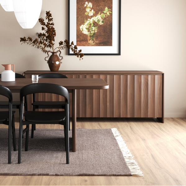 A modern dining room scene featuring a dark wood sideboard with vertical fluting, a matching wooden dining table and black chairs, a textured beige rug, and a framed floral print on the wall.