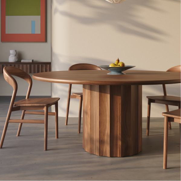 A round wooden dining table with a fluted pedestal base and matching wooden chairs in a modern dining room setting with warm beige walls and soft natural shadows.