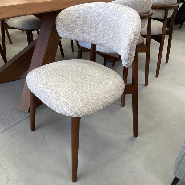 A modern dining chair with a textured light beige upholstered seat and curved backrest, supported by tapered dark brown wooden legs, shown in a dining room setting.