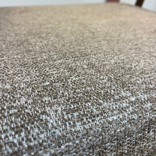 Close-up of a woven fabric upholstered surface in a warm taupe-beige color, with a blurred indoor background showing part of a chair or furniture piece.