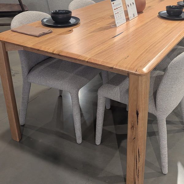 A rectangular wooden dining table with a natural oak finish, accompanied by eight light gray upholstered dining chairs, displayed in a modern furniture showroom.