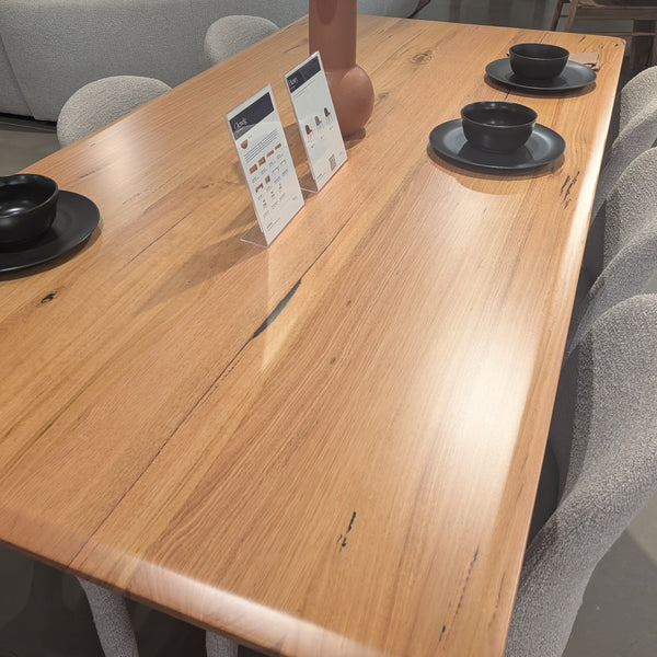 A large rectangular wooden dining table with a natural live-edge grain, styled with gray upholstered chairs and black bowls in a modern dining room showroom.