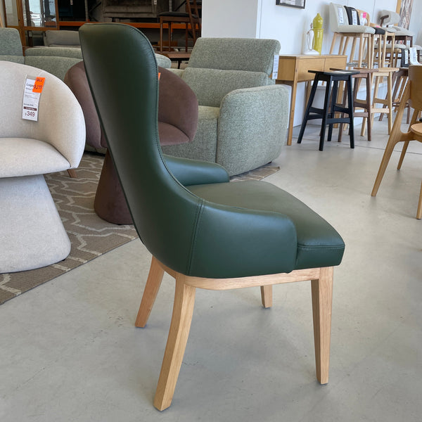A modern dining chair with a dark green upholstered seat and backrest, supported by light natural wood legs, shown in a furniture showroom.