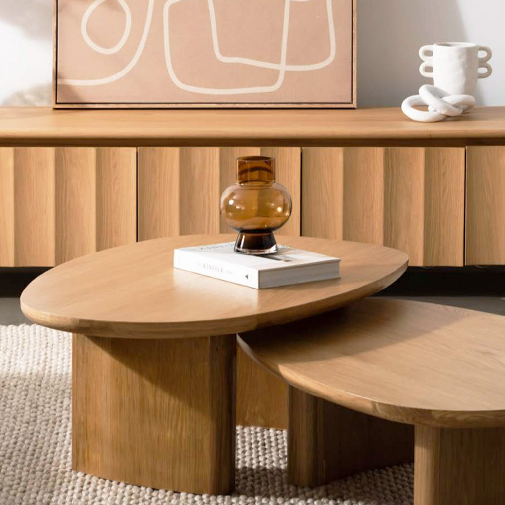 A set of three nested oval nesting coffee tables in a warm light oak wood finish, styled with a small amber glass vase and a white book in the center, with a matching wooden credenza in the background.