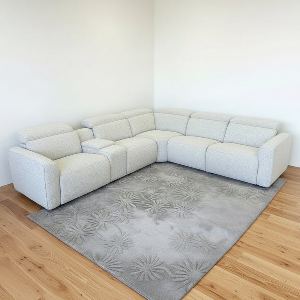 A modern curved sectional sofa in a light beige textured fabric, arranged in a showroom with matching armchairs, three round bar stools with reddish-brown cushions, and a patterned area rug.