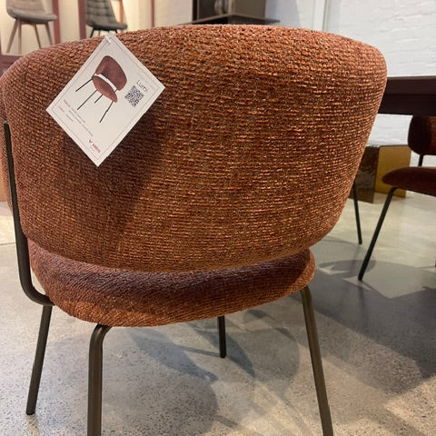 A close-up of a modern upholstered accent chair with a rounded, high-back curved backrest in a textured rust-brown bouclé fabric, supported by slim black metal legs. A white product tag is attached to the backrest, and the background shows a showroom interior with other furniture pieces.