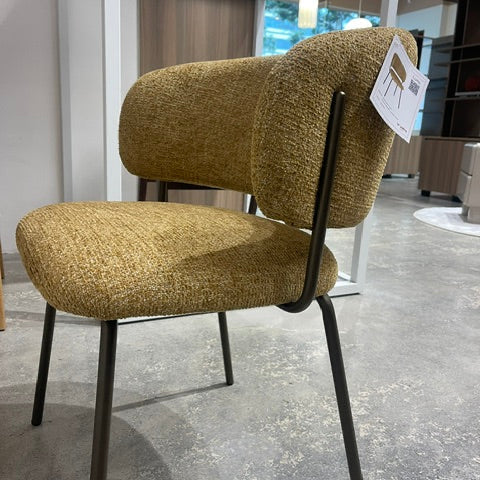 A modern upholstered dining chair with a textured mustard-yellow bouclé seat and curved backrest, supported by a black metal frame, shown in a furniture showroom.