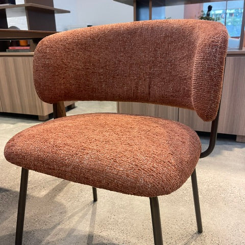 A modern upholstered dining chair with a textured terracotta orange-brown fabric on the seat and curved backrest, supported by slim black metal legs, shown in a showroom setting.