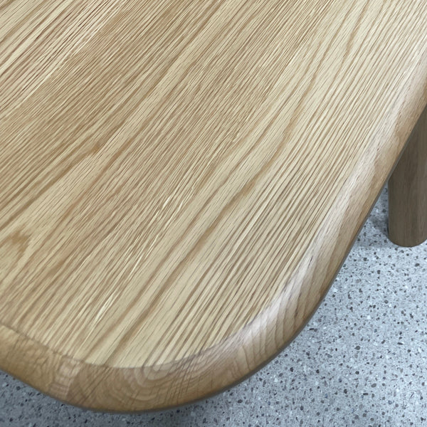 Close-up of a light oak wood table corner with rounded edges and visible wood grain, resting on a speckled gray floor.