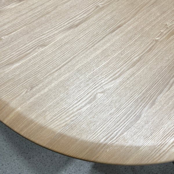Close-up of a light oak wood tabletop with visible grain, showing the curved edge and part of the table base over a speckled gray floor.