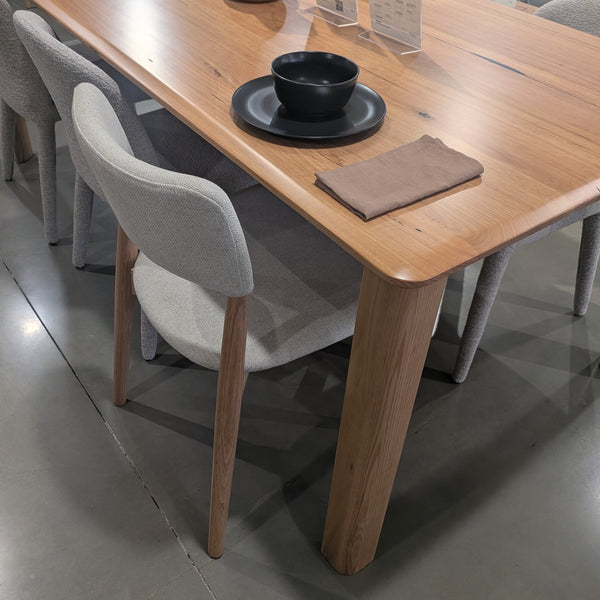 A rectangular wooden dining table with a natural oak finish, accompanied by light gray upholstered chairs, shown in a furniture showroom.