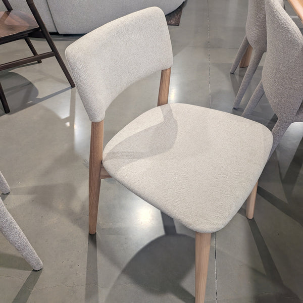 A modern dining chair with a light beige textured fabric seat and backrest, supported by light natural wood legs, shown in a furniture showroom.