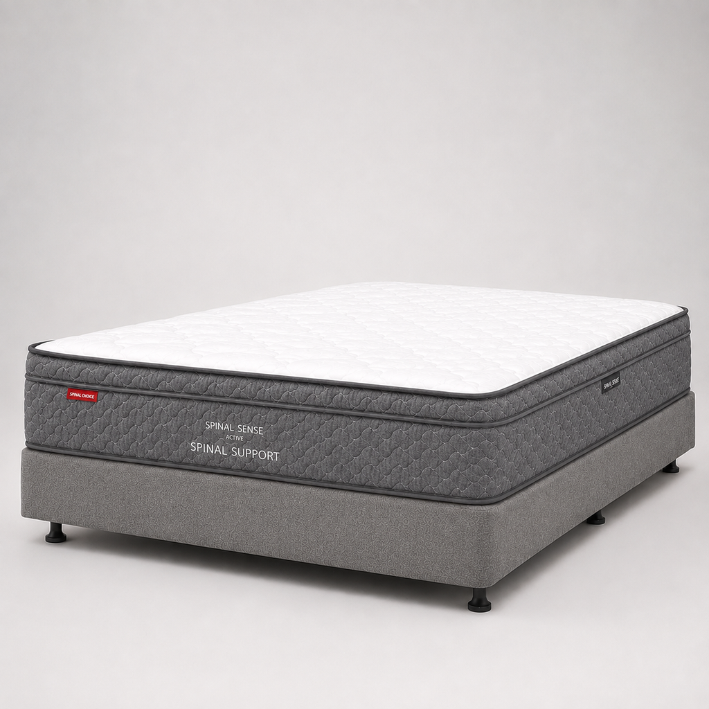 A mattress on a gray upholstered support base, photographed in a clean studio setting with a white background. The mattress has a white quilted top and dark gray sides with visible text reading 'SPINAL SUPPORT' and a small red logo tag.